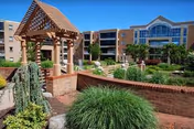 Outdoor courtyard area of a senior living facility with a wooden pergola, brick walls, green shrubs, and a multi-story brick building in the background under a clear blue sky.