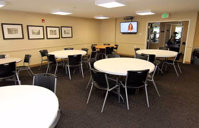 A room with multiple round tables covered with white tablecloths, each surrounded by black chairs. The walls are beige with framed artwork hanging on one side. A flat-screen TV is mounted on the far wall, and an open doorway leads to another room with exercise equipment visible.