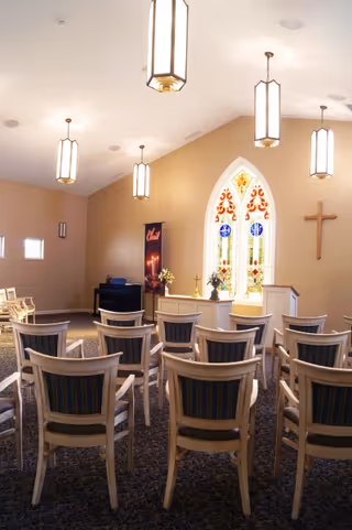 Small chapel-style room with rows of chairs facing a stained-glass window and a wooden cross.