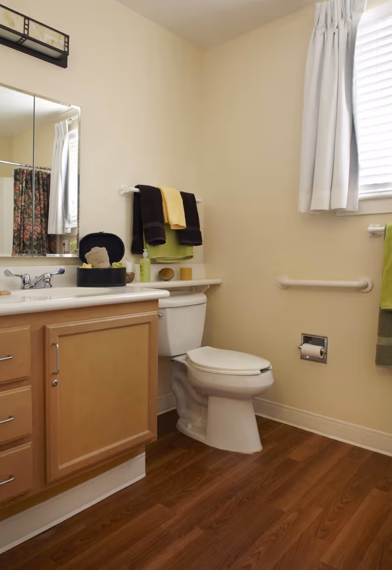 A clean bathroom with a wooden vanity cabinet, a white sink, a toilet, and a large mirror above the sink. There are towels hanging on a rack above the toilet and a window with white curtains letting in natural light. The floor is wood-style laminate, and there are grab bars on the walls near the toilet.