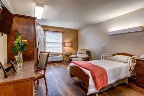 A furnished senior living facility bedroom with a bed, dresser, chair, and window.