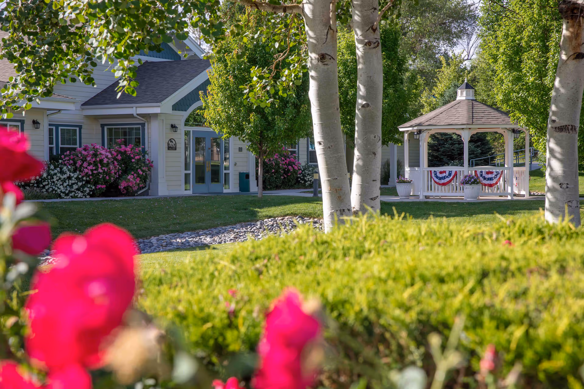 A peaceful outdoor garden area at Crossroads at Delta featuring a white gazebo decorated with red, white, and blue bunting. The scene includes green trees, bushes, and colorful flowers with a building entrance visible in the background.