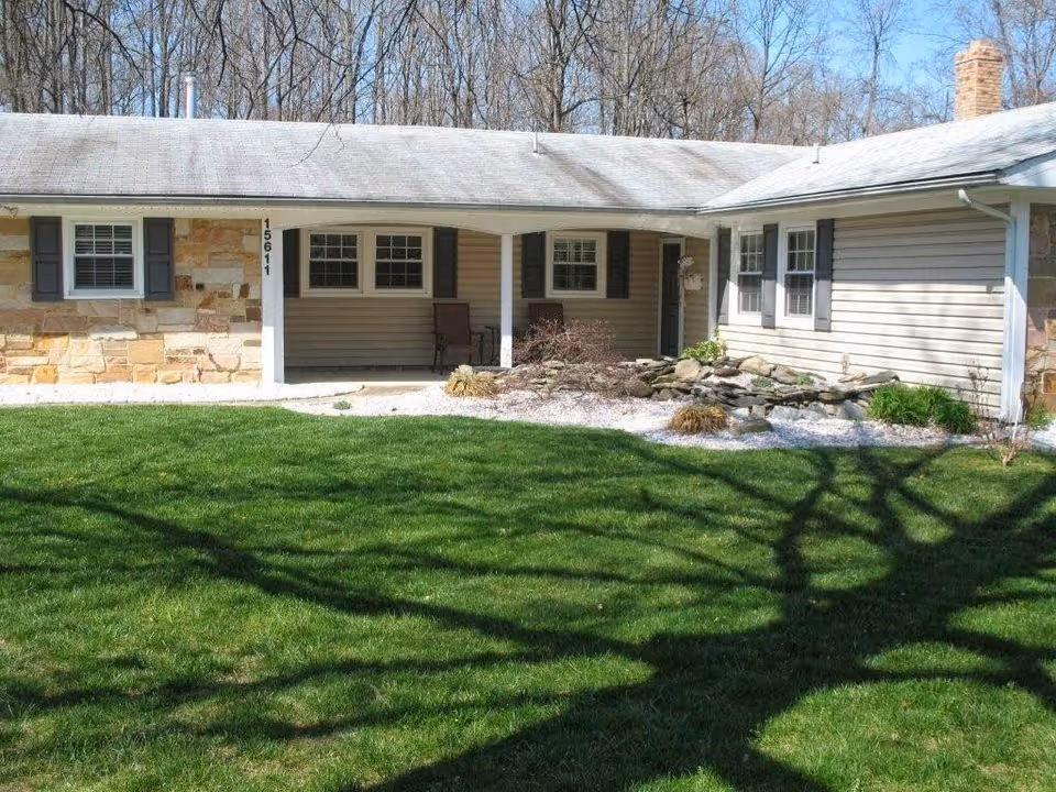 Single-story building with a covered porch area, stone and siding exterior, several windows with shutters, a small rock garden, and a well-maintained green lawn in front. Trees without leaves are visible in the background.