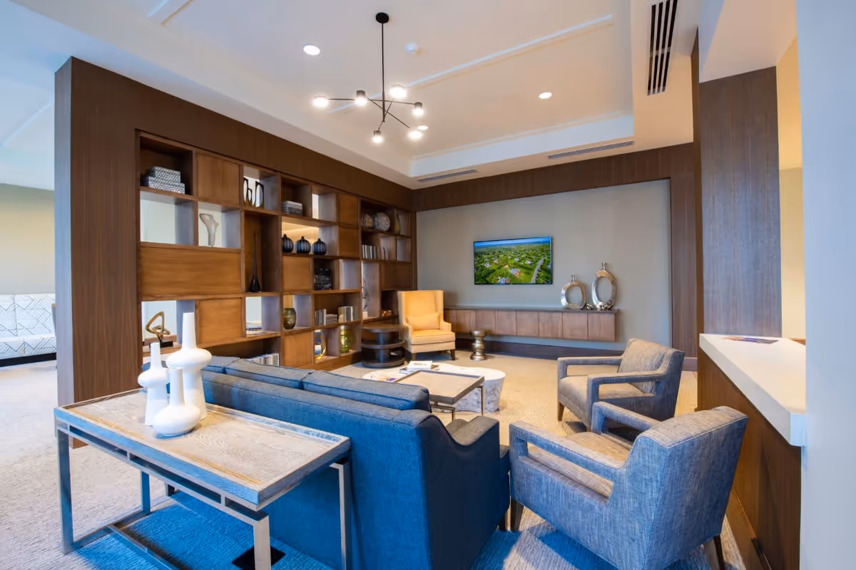 A modern living room area with a blue sofa, two blue armchairs, a yellow armchair, a wooden coffee table, and a wooden shelving unit with decorative items. A flat-screen TV is mounted on the wall above a floating wooden cabinet. The room has a contemporary chandelier and neutral-colored walls and carpet.