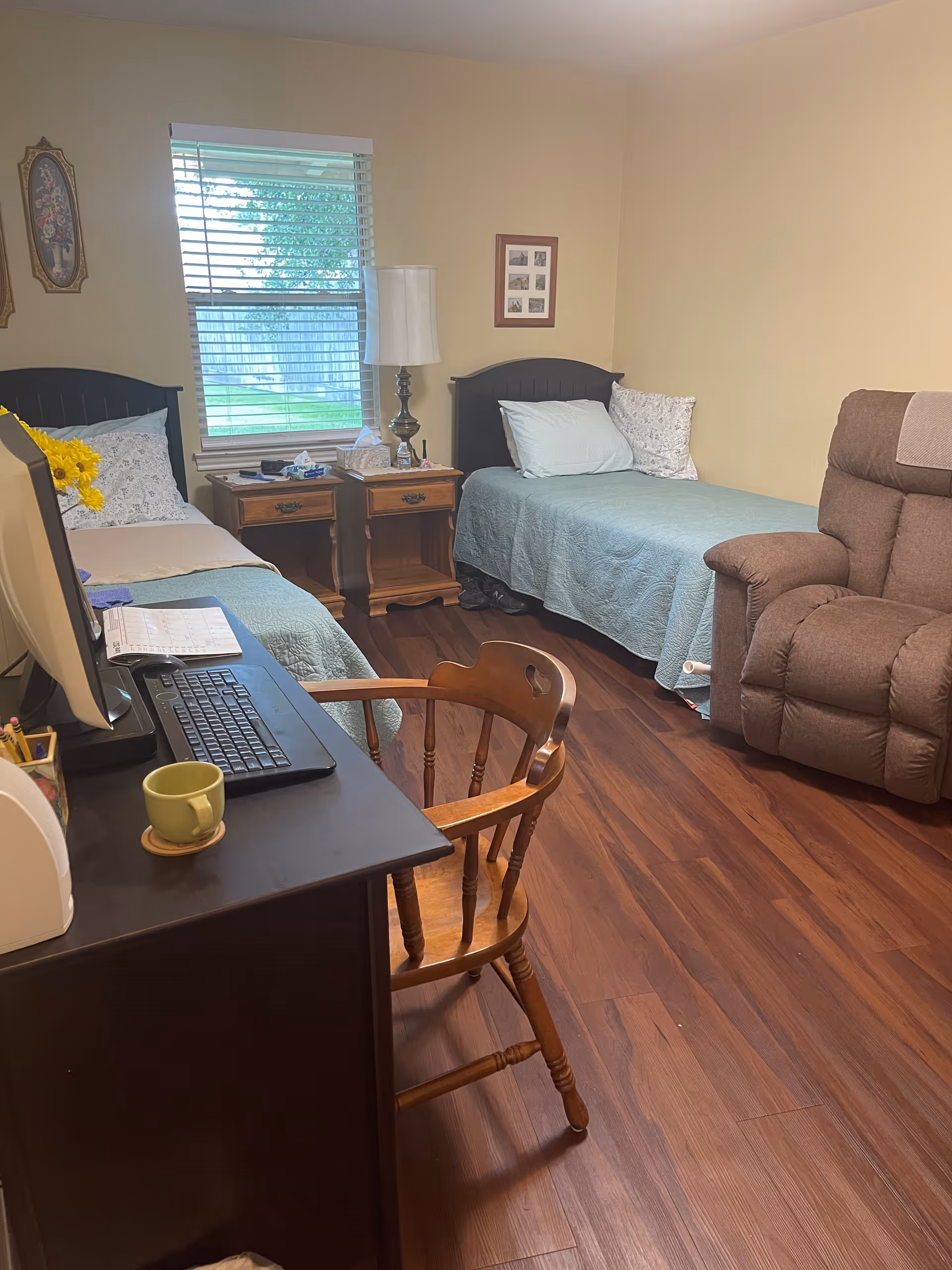 A cozy room with two twin beds covered in light blue quilts, a wooden desk with a computer and keyboard, a wooden chair, two wooden nightstands with a lamp and tissues, a brown recliner chair, and a window with blinds showing a green yard outside.
