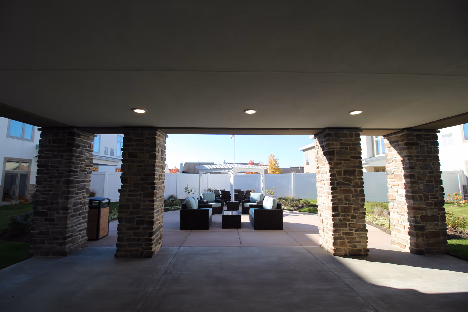 Outdoor seating area with cushioned chairs and a table under a covered patio supported by stone pillars, with a white fence and buildings in the background.