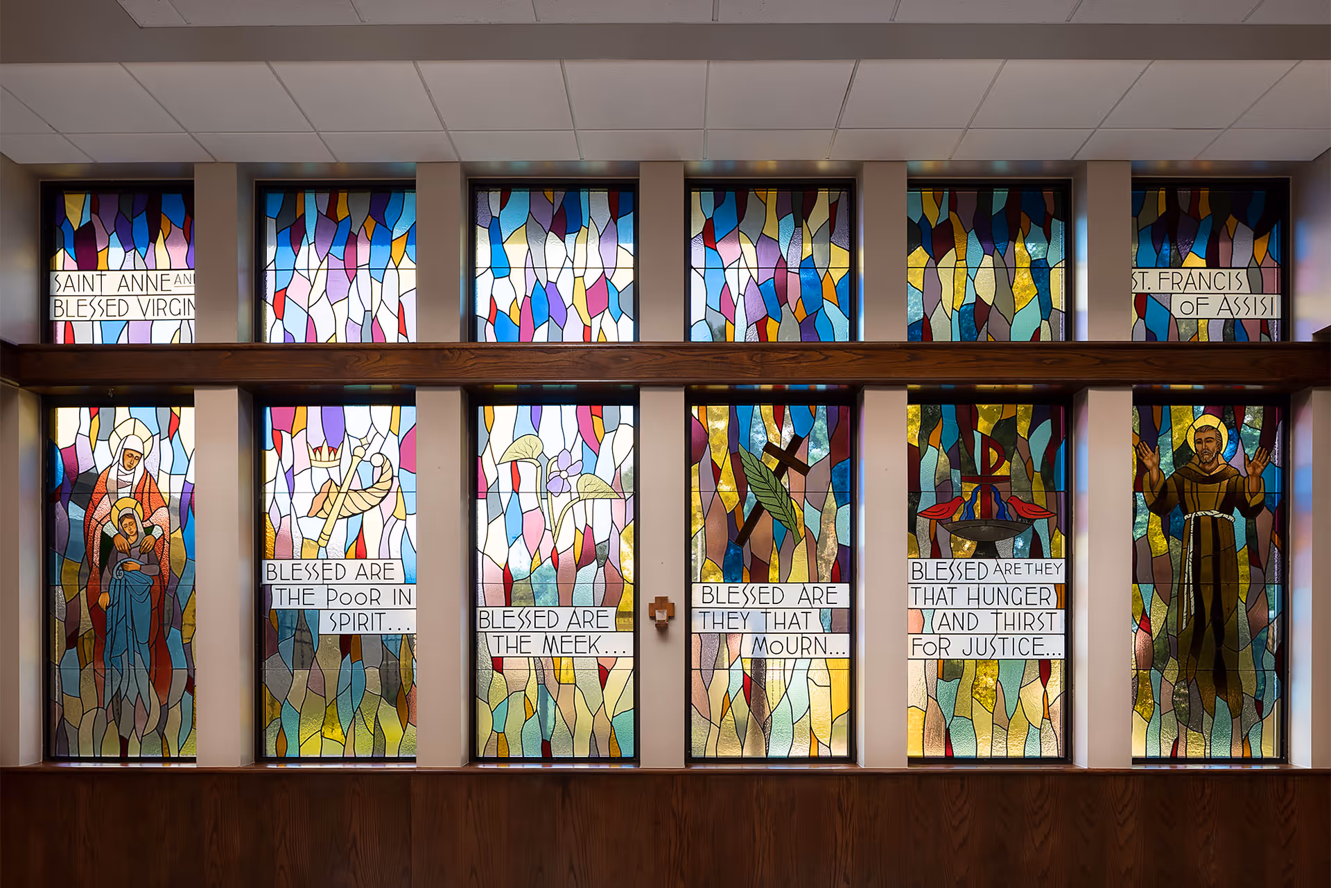 A row of colorful stained-glass chapel windows depicting religious figures and panels with Beatitudes text.