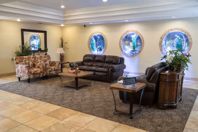 A cozy seating area in a senior living facility with a brown leather sofa, two floral patterned armchairs, a wooden coffee table, and side tables with plants and a lamp. The walls are light-colored with three round windows decorated with blue and green glass art. The floor is tiled with a large patterned rug under the furniture.