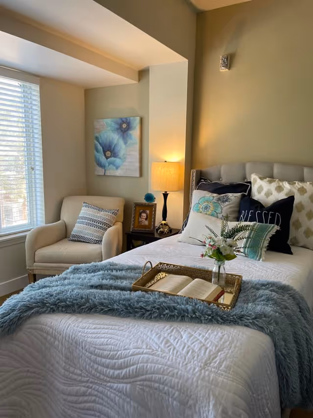 A cozy bedroom with a neatly made bed featuring multiple decorative pillows and a blue throw blanket. A gold tray on the bed holds an open book and a small vase with flowers. Next to the bed is a nightstand with a lamp, a framed photo, and a decorative item. A comfortable armchair with a patterned pillow sits near a window with white blinds. A painting of blue flowers hangs on the wall above the chair.