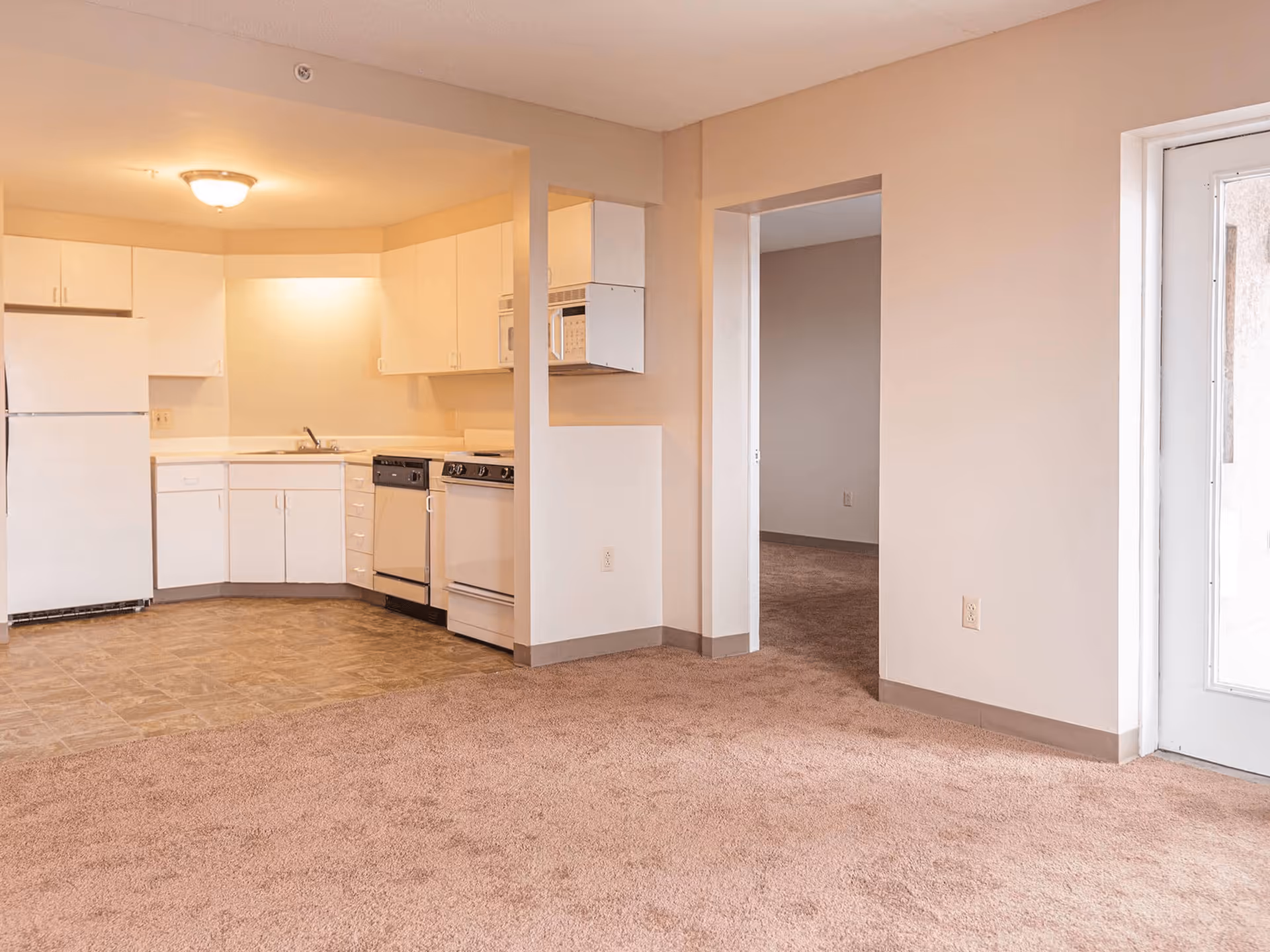 Interior view of an apartment showing a small kitchen area with white cabinets, a refrigerator, dishwasher, stove, and microwave. The kitchen floor is tiled, and the adjacent living area is carpeted. There is an open doorway leading to another room and a glass door letting in natural light.