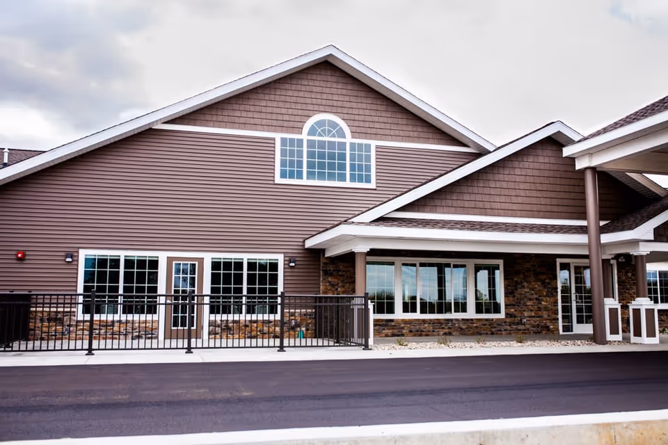 Front exterior of a brown-clad senior living building with large windows, a covered entrance, and a paved driveway.