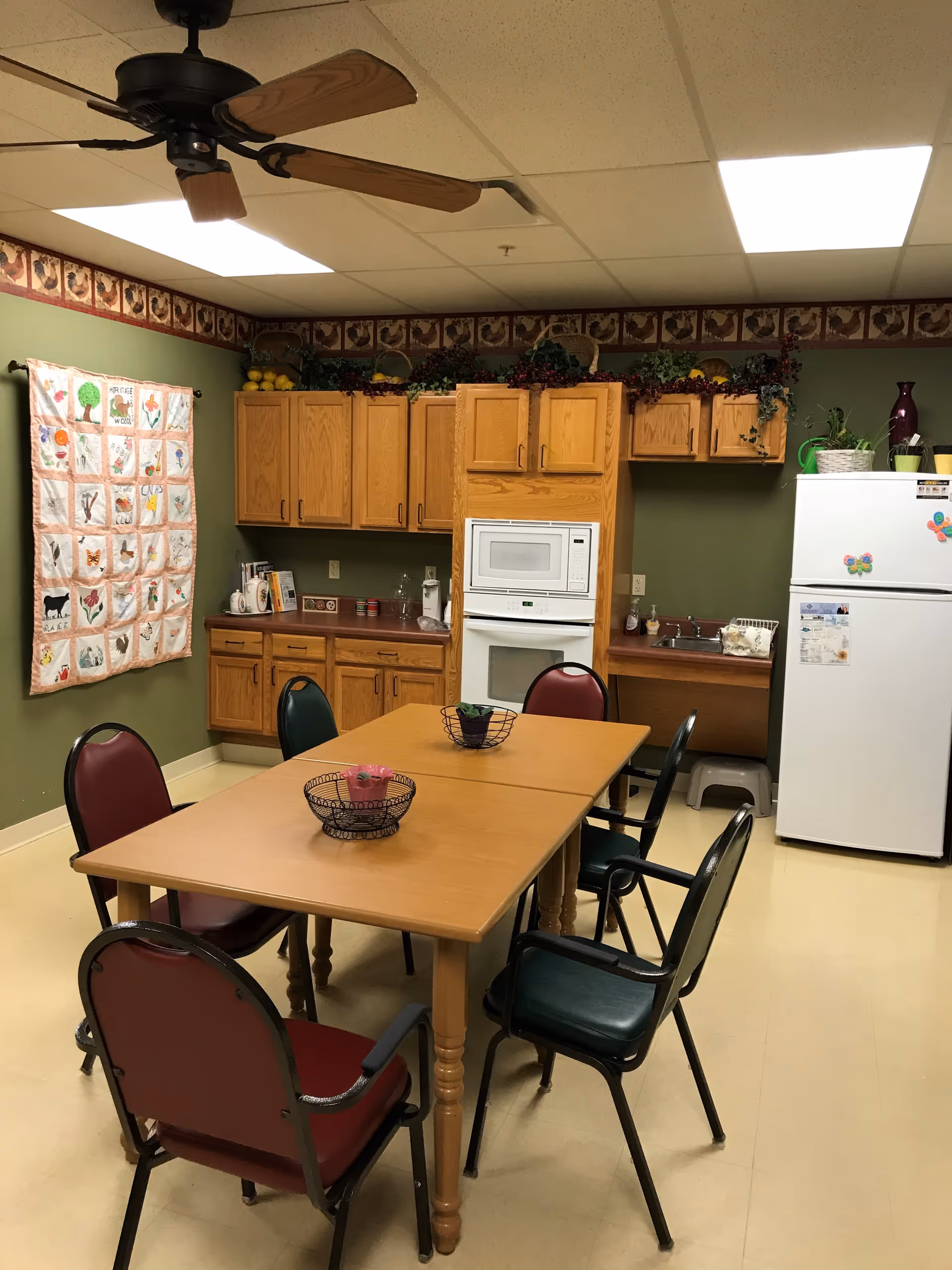 A small kitchen and dining area with a wooden table surrounded by six chairs, a ceiling fan above, wooden cabinets, a white refrigerator decorated with colorful magnets, a microwave, and a sink. The walls are green with a decorative border near the ceiling and a quilt hanging on one wall.