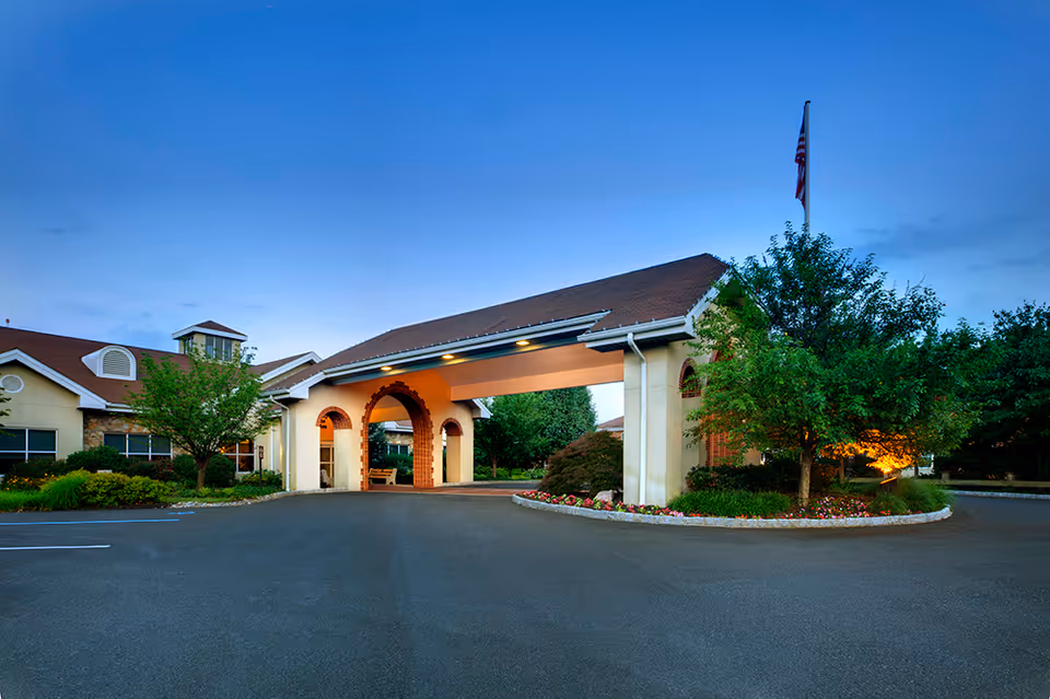 Covered entrance/porte-cochère of a senior living facility with landscaped beds, trees, and an American flag.