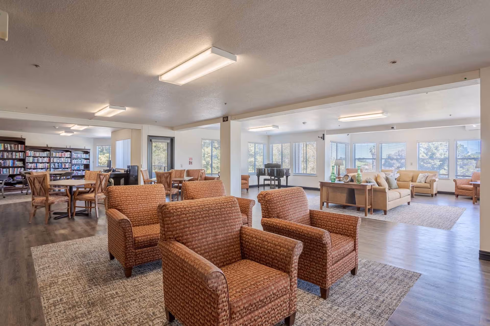 A spacious senior living common area with multiple seating arrangements including patterned armchairs and beige sofas. The room features large windows allowing natural light, a piano in the corner, bookshelves filled with books, and several tables with chairs. The flooring is a combination of wood and area rugs, and the ceiling has rectangular fluorescent lights.