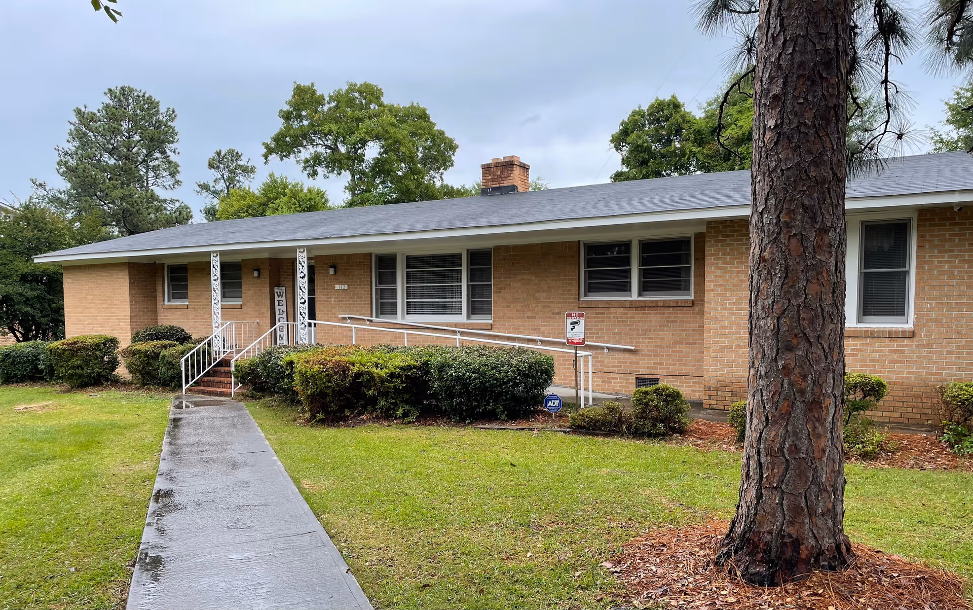 Single-story brick residential building with a front walkway, wheelchair ramp, shrubs and a large tree in the yard.