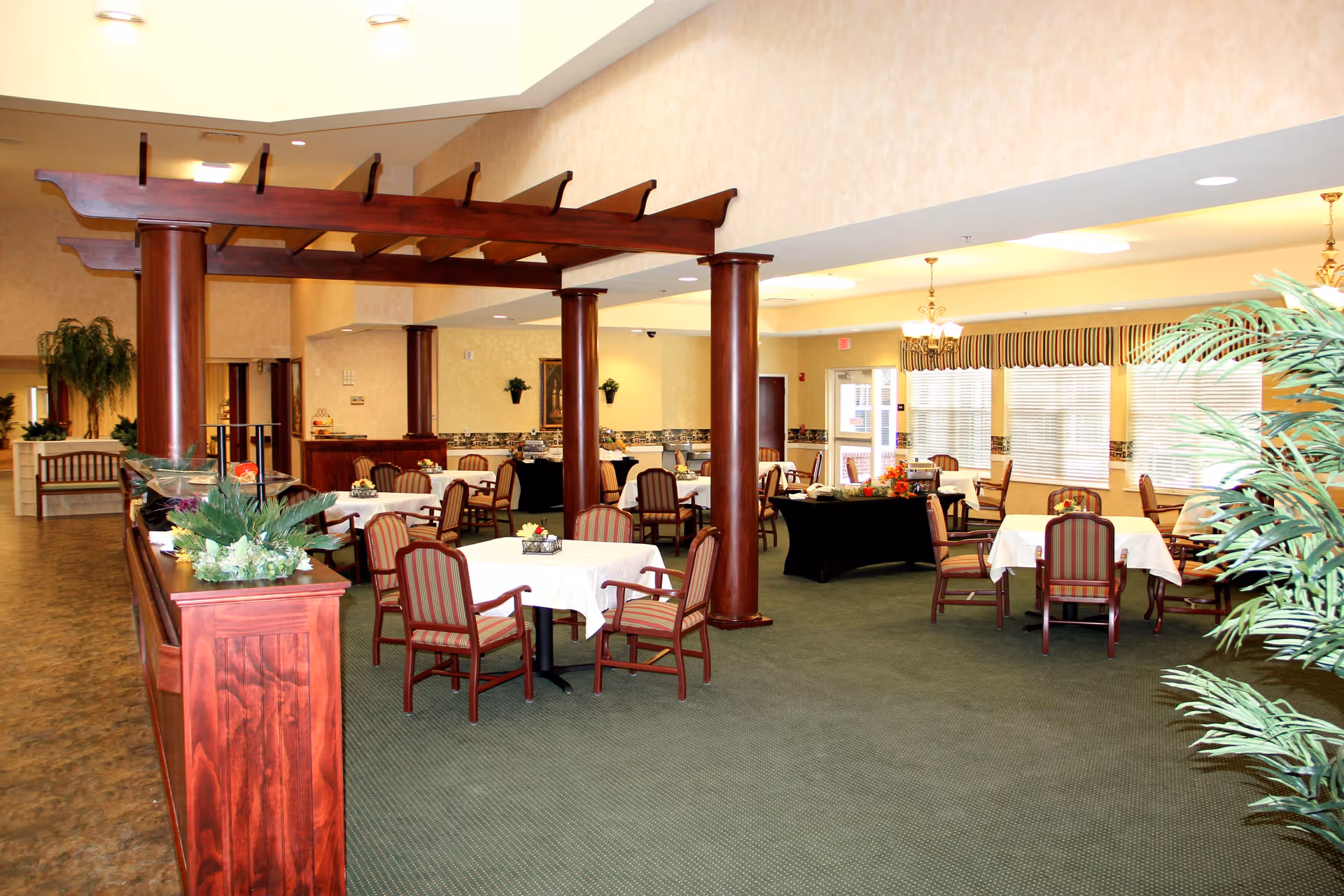 A spacious dining room with multiple tables covered with white tablecloths and surrounded by wooden chairs with striped cushions. The room features wooden columns and a decorative wooden pergola structure. Large windows with striped valances allow natural light to fill the space. There are plants and floral arrangements adding greenery and color to the room.