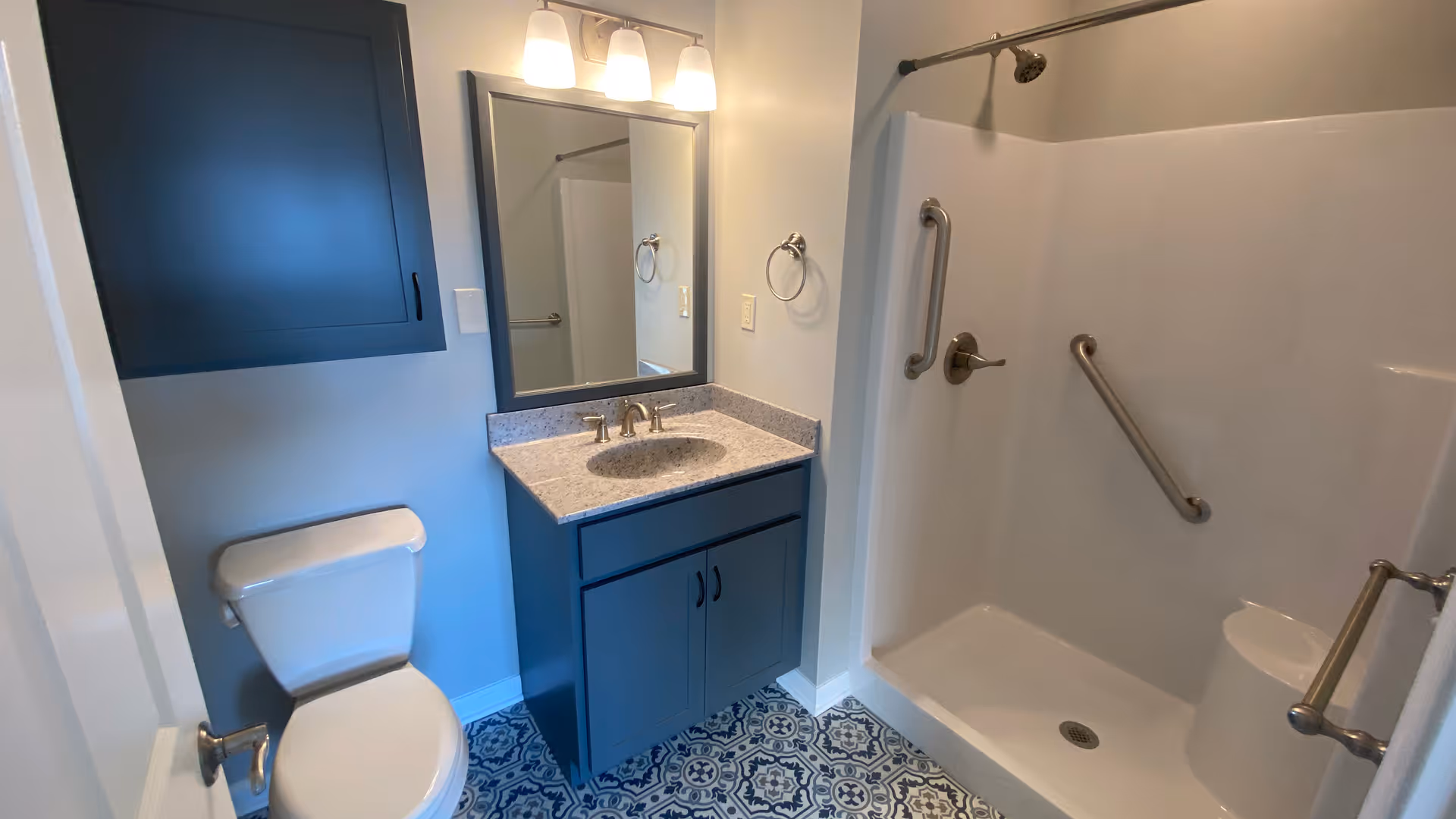 A clean bathroom featuring a white toilet, a blue vanity with a granite countertop and sink, a large mirror with three overhead lights, a blue wall cabinet, and a walk-in shower with multiple grab bars and a built-in seat.