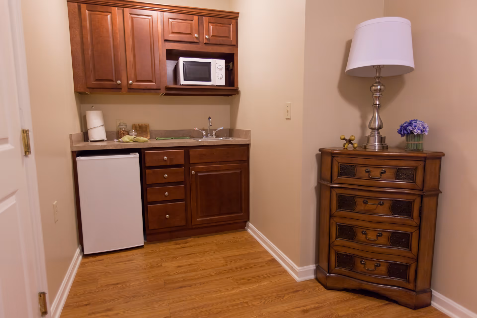 Compact kitchenette with cabinets, microwave, mini-fridge, sink, and nearby dresser.
