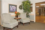 Two patterned armchairs with a small round wooden table between them holding a vase with flowers. A framed floral picture hangs on the wall above the chairs, and a tall green potted plant is placed near the doorway leading to a hallway with wooden flooring and visible ceiling lights.