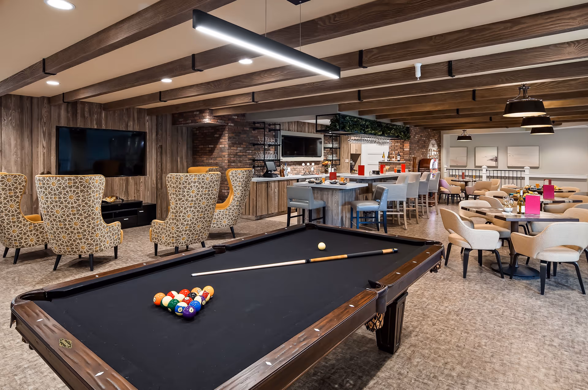 A communal lounge showing a pool table in the foreground with seating areas, dining tables, and a bar in the background.
