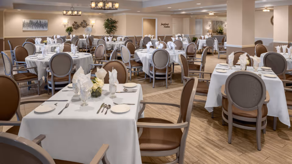A spacious dining room in a senior living facility with multiple round and rectangular tables covered with white tablecloths. Each table is set with plates, silverware, glasses, and folded napkins. The room features comfortable chairs with armrests, soft lighting fixtures, and decorative plants and artwork on the walls.