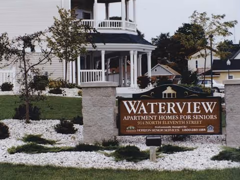 Exterior view of Waterview Apartment Homes for Seniors, showing a landscaped area with white rocks, small trees, and a sign displaying the facility's name and address in front of a white building with a porch.