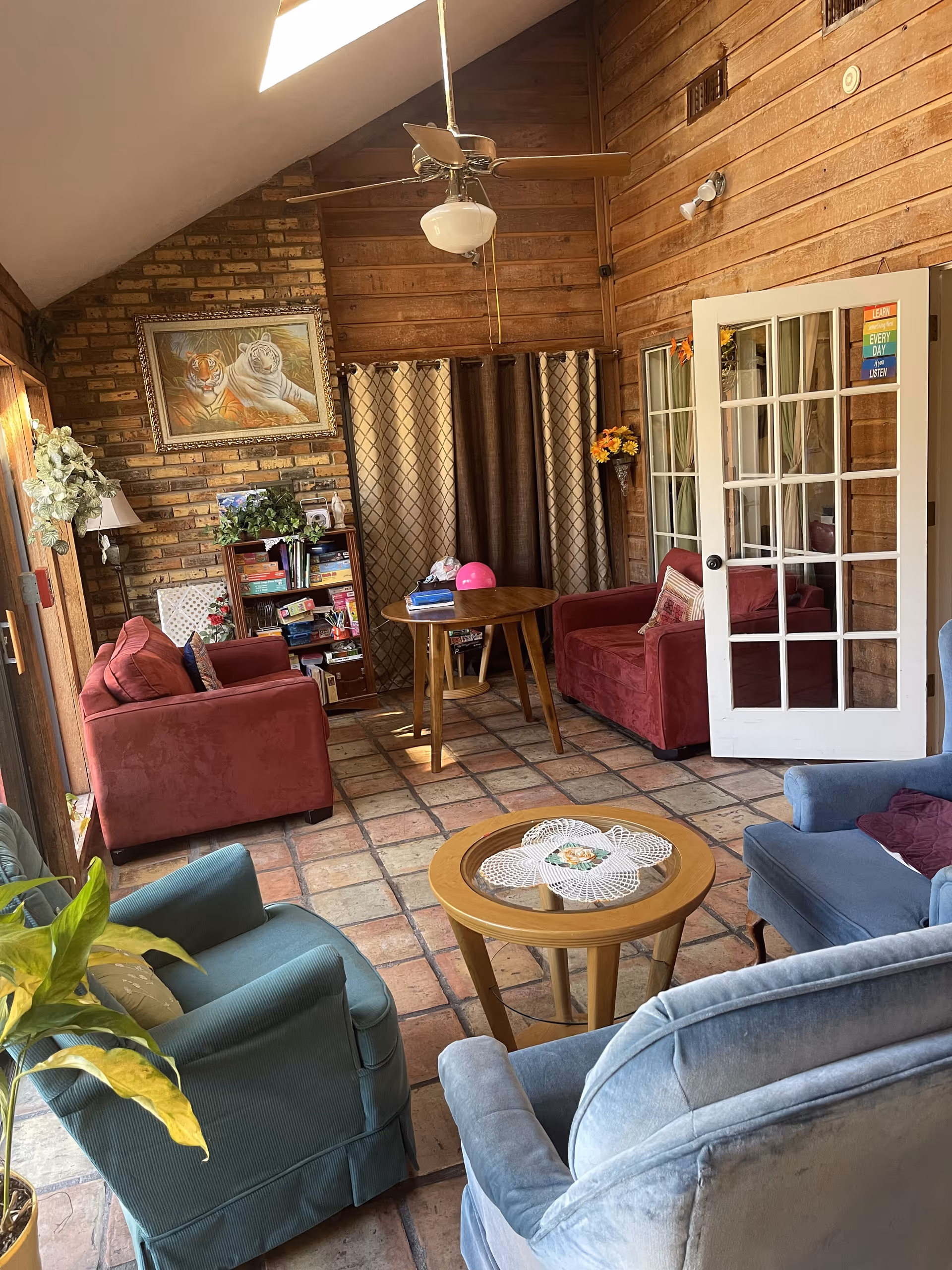 A cozy living room with wooden walls and a tiled floor. The room features two red sofas, two blue armchairs, a round wooden coffee table with a doily, and a small round dining table with books and a pink helmet on it. A ceiling fan with a light hangs from the vaulted ceiling. There is a bookshelf filled with books and plants, a framed painting of two tigers on the brick wall, and a glass-paneled door with colorful motivational stickers.