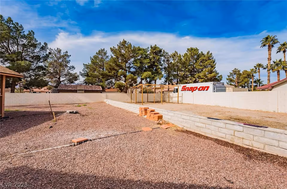 Outdoor area with gravel ground, a low retaining wall, some stacked bricks, and a wooden pergola frame. The area is enclosed by a white block wall with trees and palm trees visible beyond the wall under a partly cloudy blue sky.