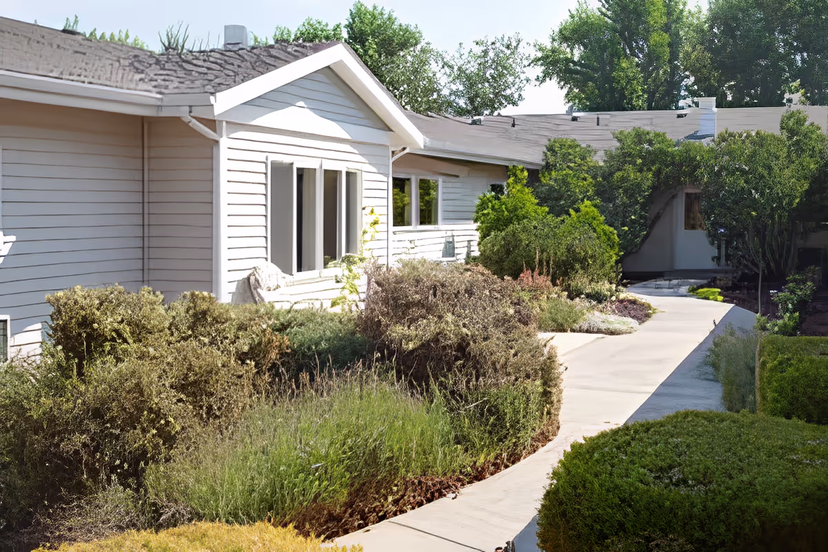 Paved walkway curves past landscaped shrubs to the front of a single-story white-sided senior living building.