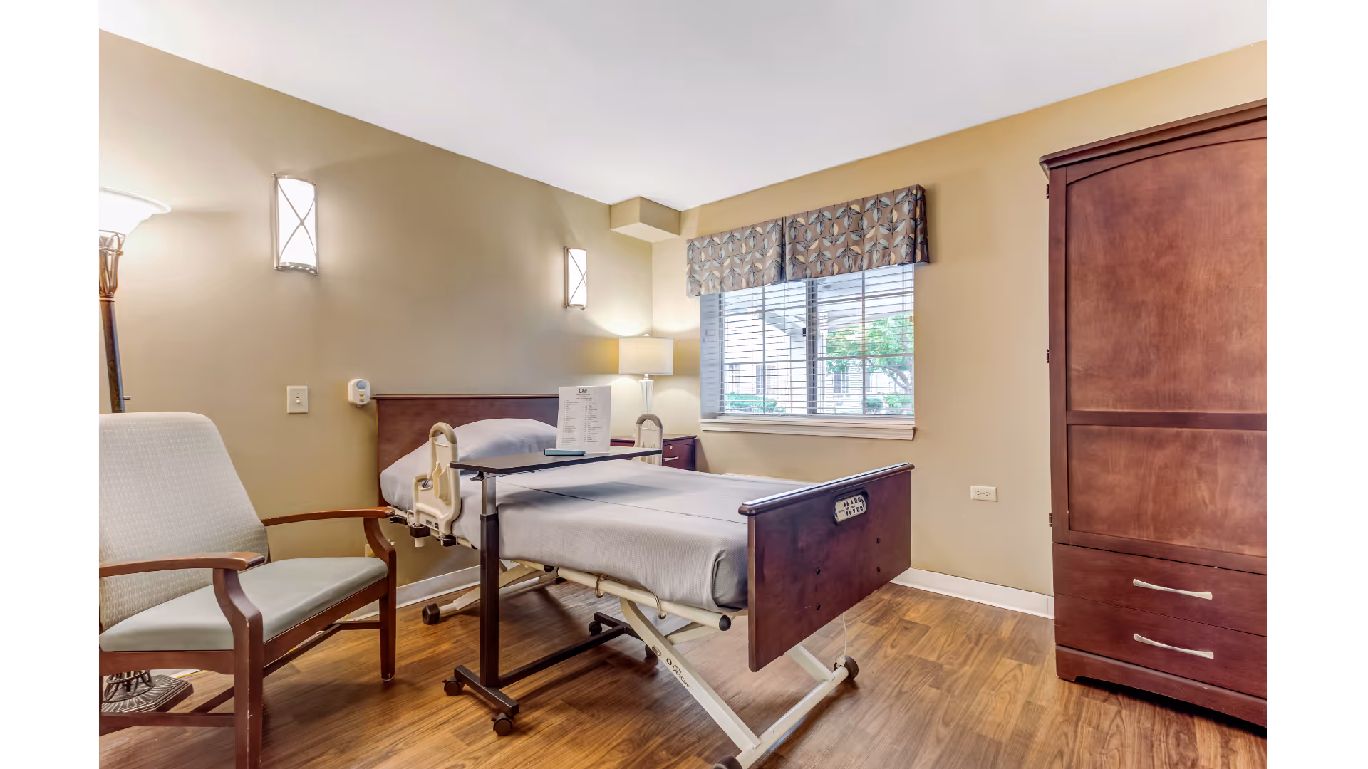 A furnished assisted-living bedroom with a hospital-style bed, armchair, window, and wooden armoire.