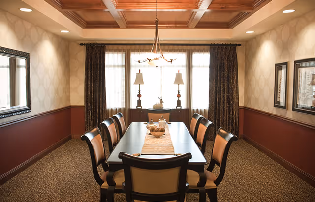 A formal dining room with a long rectangular table surrounded by eight chairs. The table has a decorative runner with a bowl and glass jars on it. The room features patterned wallpaper, a coffered wooden ceiling, carpeted floor, and large windows with curtains letting in natural light. Two table lamps and framed artwork are visible in the background.