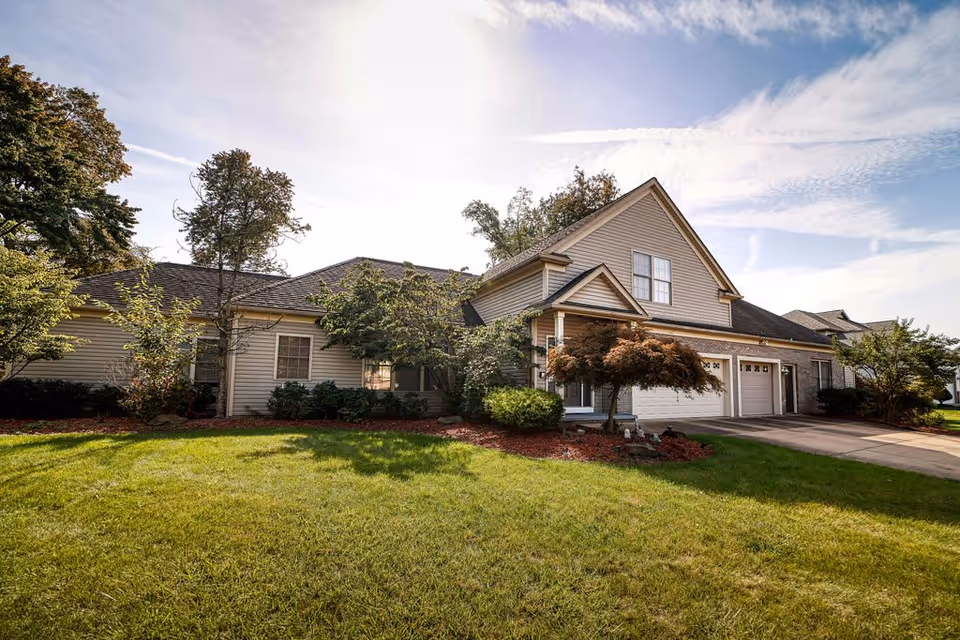 Exterior view of a residential-style building with beige siding, multiple windows, and a three-car garage. The building is surrounded by green grass, trees, and shrubs under a partly cloudy sky.
