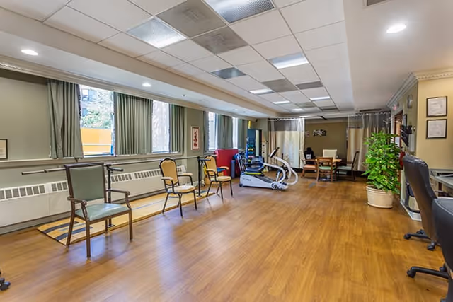 A spacious room with wooden flooring and several chairs arranged along the walls. The room has large windows with green curtains, exercise equipment including a stationary bike, a table with chairs in the back, and a potted plant near a desk with a computer chair.