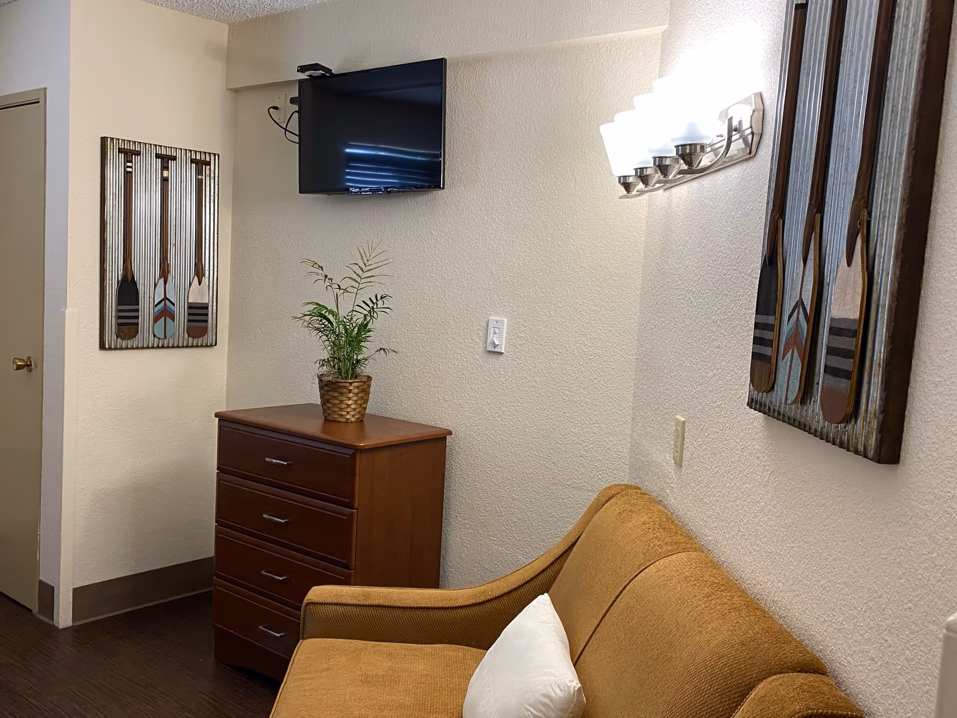 A small living area with a brown couch featuring a white pillow, a wooden chest of drawers with a potted plant on top, a wall-mounted flat screen TV, and decorative wall art of three wooden paddles. The walls are light-colored and there is a modern wall light fixture above the couch.