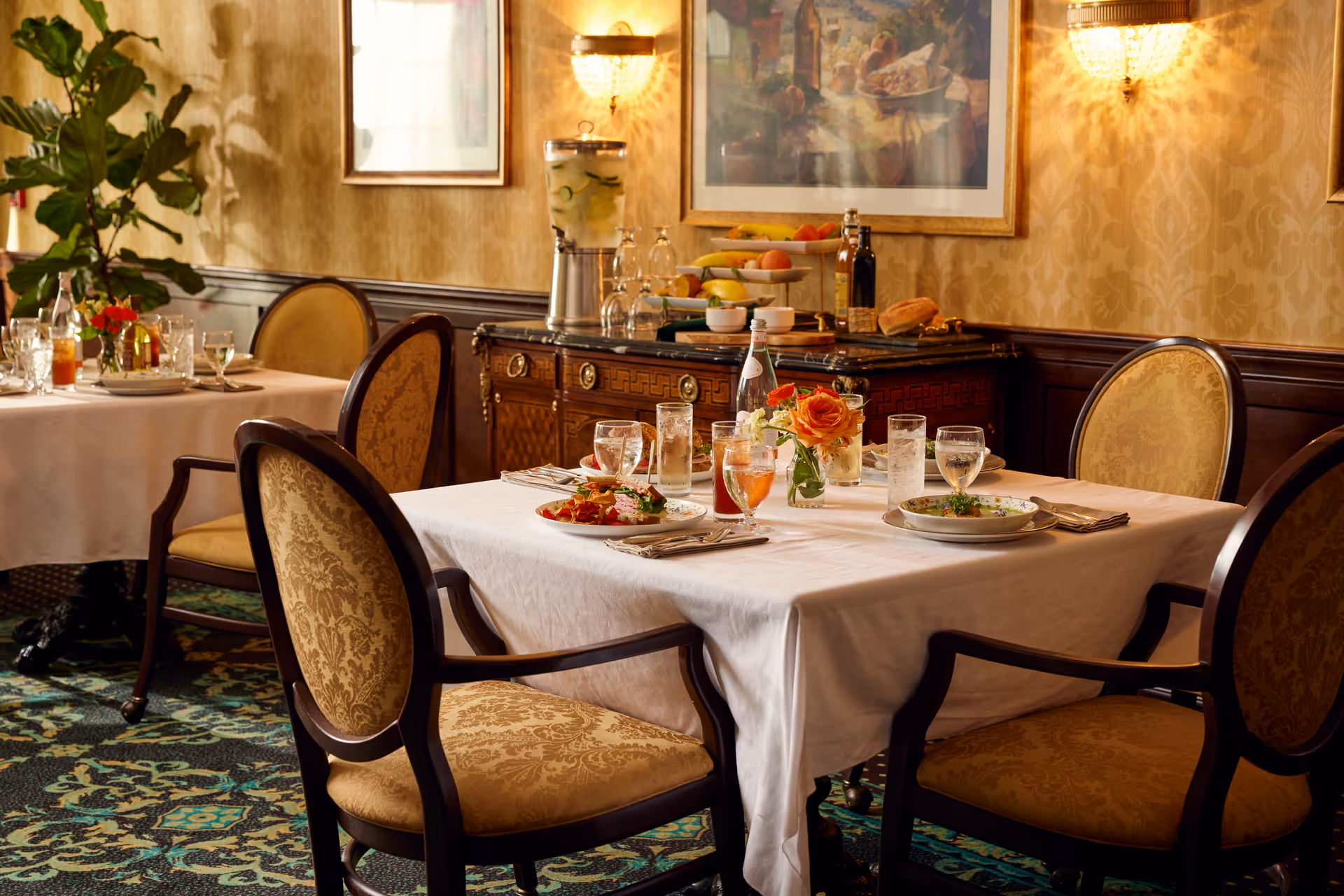 A dining room with a table set for four, featuring plates of food, glasses of water and iced tea, and a small vase with an orange rose. The room has elegant upholstered chairs, patterned carpet, wallpaper with a classic design, and a sideboard with a water dispenser, fruit, and condiments. Warm lighting fixtures and framed artwork adorn the walls.