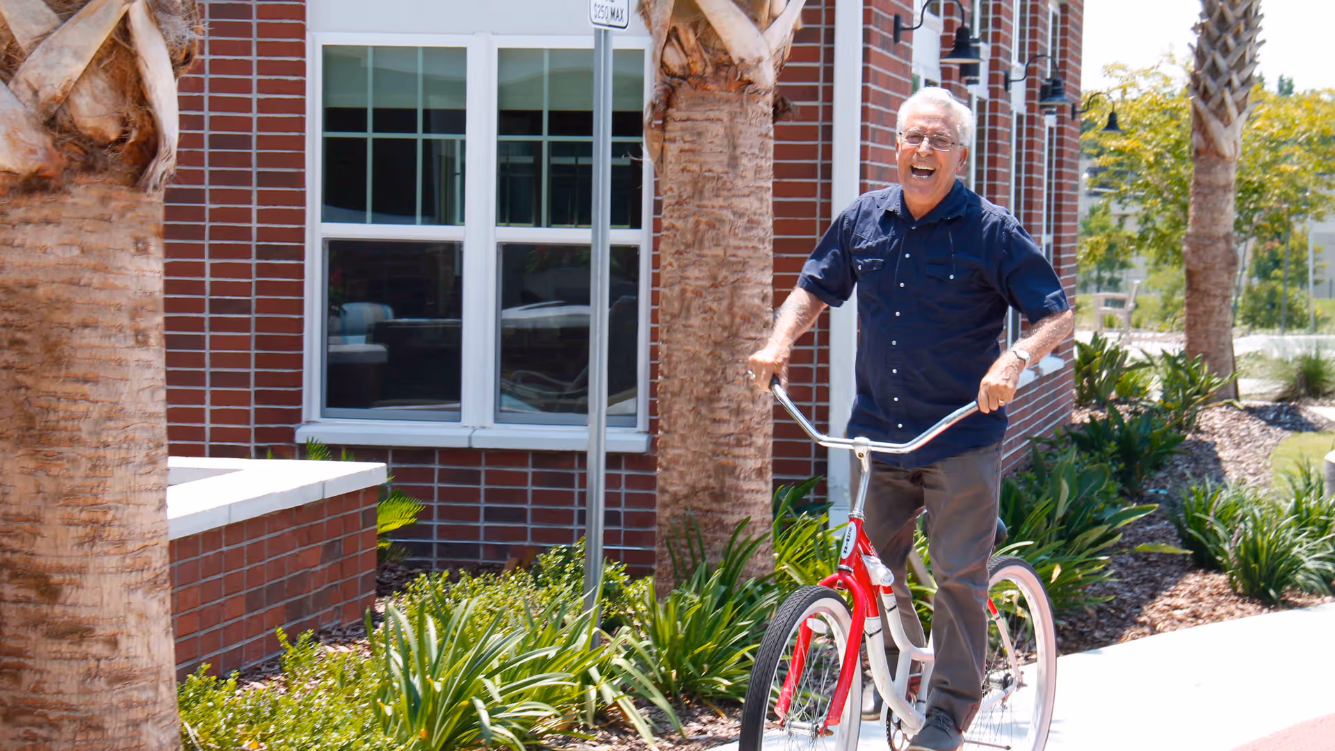 An elderly man smiling and riding a red and white bicycle on a sidewalk next to a brick building with windows and palm trees, surrounded by green plants and landscaping.