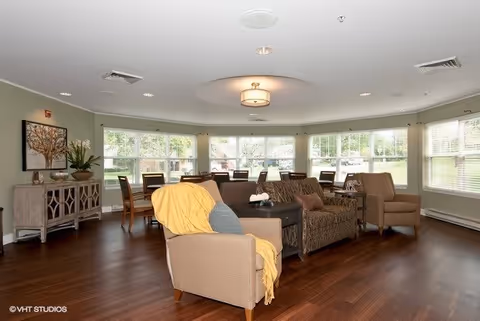 A spacious living room area with multiple armchairs and sofas arranged around a central coffee table. Large windows line the curved wall, allowing natural light to fill the room. The floor is wooden, and there is a sideboard with decorative items and a framed artwork on the wall. The ceiling has recessed lighting and a central light fixture.