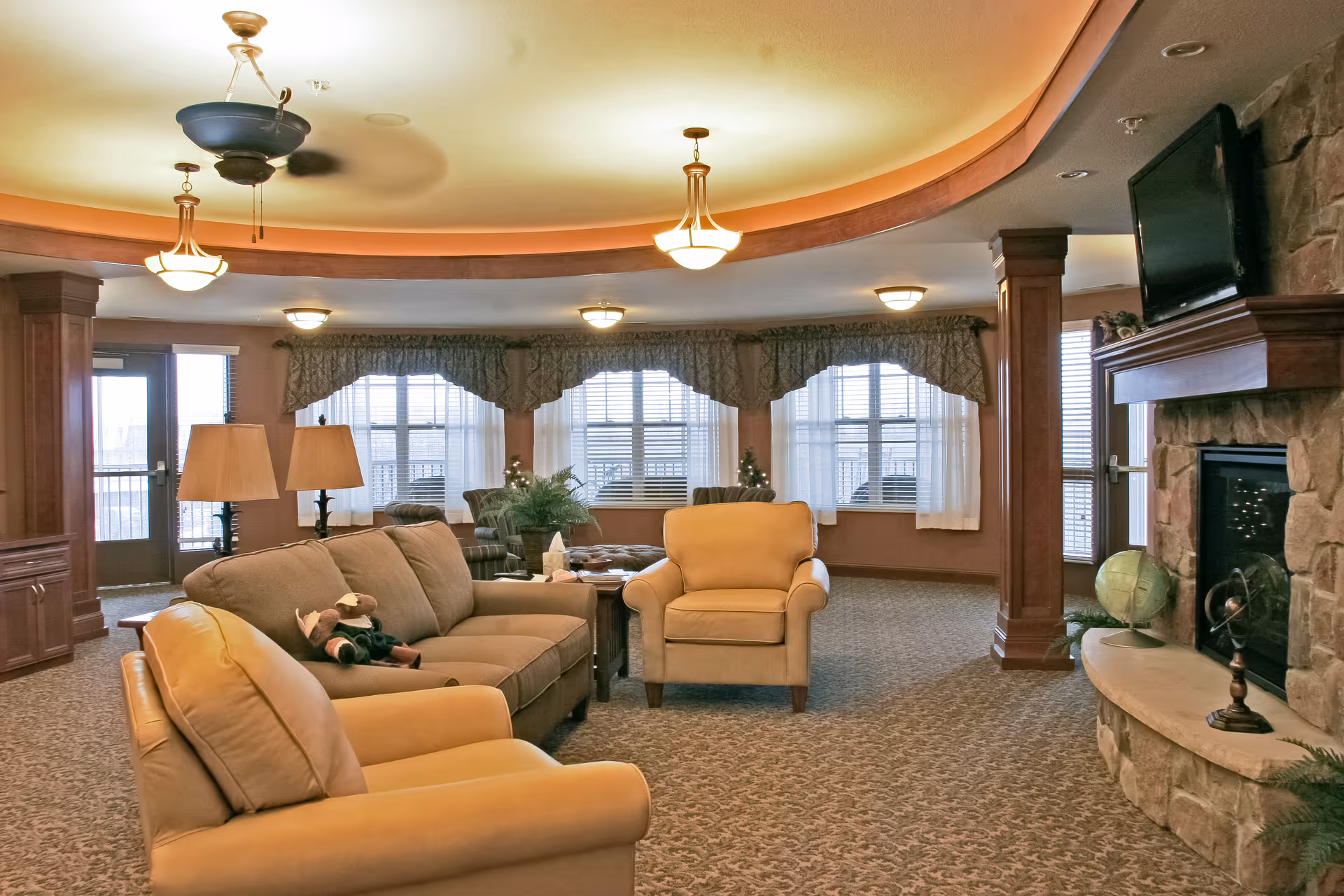 A cozy senior living facility common area with beige and brown upholstered armchairs and a sofa arranged around a stone fireplace with a mounted TV above it. The room has large windows with valances and sheer curtains, carpeted floors, and warm lighting from ceiling fixtures and table lamps. There are decorative plants and a globe on the fireplace mantel.