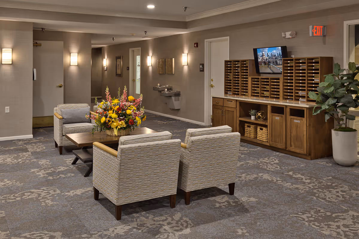 A seating area in a senior living facility with four cushioned armchairs arranged around a wooden coffee table that holds a large colorful flower arrangement. The room has patterned carpet, beige walls with wall-mounted lights, a water fountain, and a wooden cabinet with multiple mail slots and a small TV mounted above it. There is also a large potted plant near the cabinet and an exit sign above a door in the background.