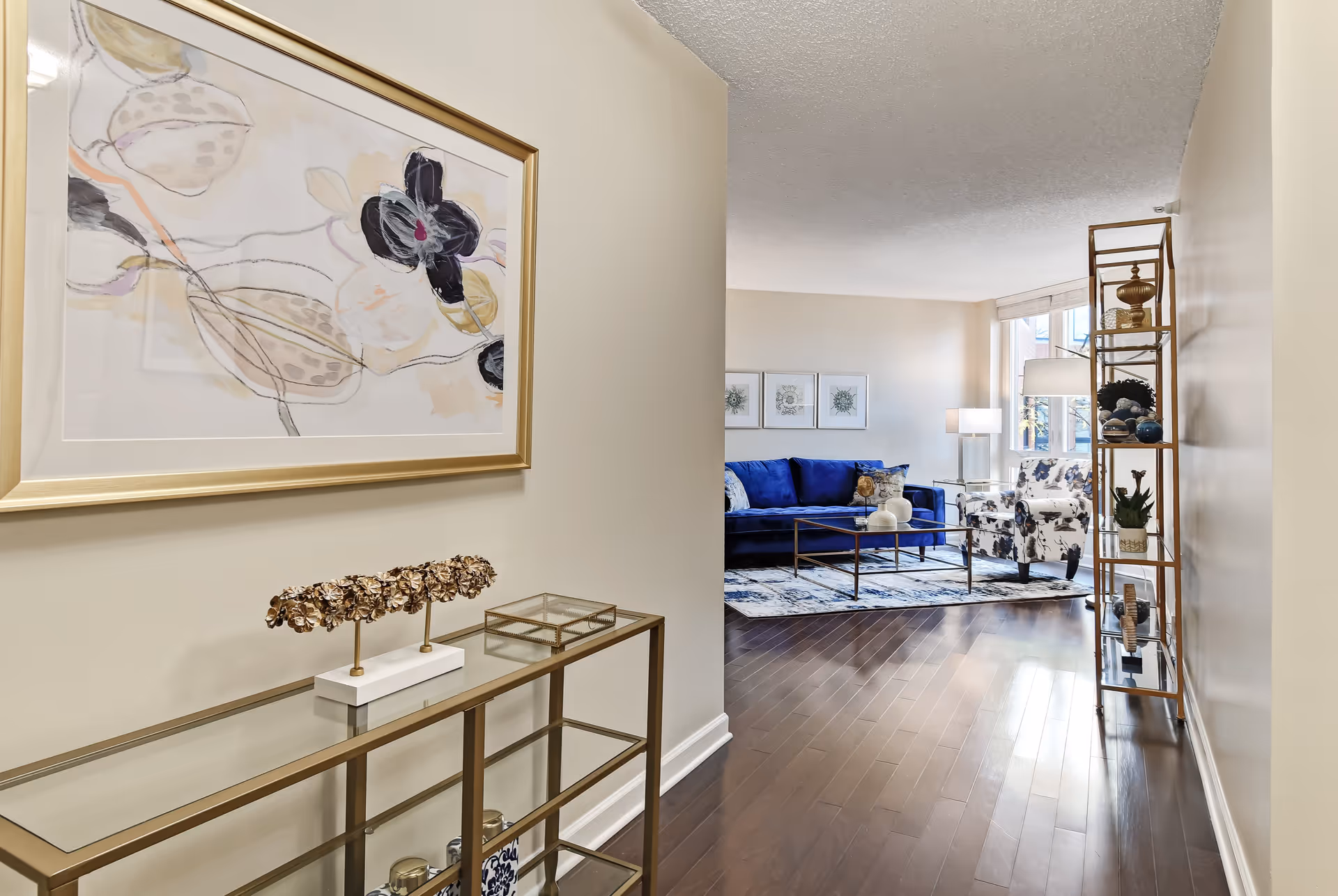 View down a hallway with a glass and gold metal console table on the left holding decorative items and a framed abstract floral painting above it. At the end of the hallway is a living room with a blue sofa, a floral armchair, a glass coffee table, a floor lamp, and a gold metal shelving unit with various decor items. The room has wooden floors and large windows letting in natural light.