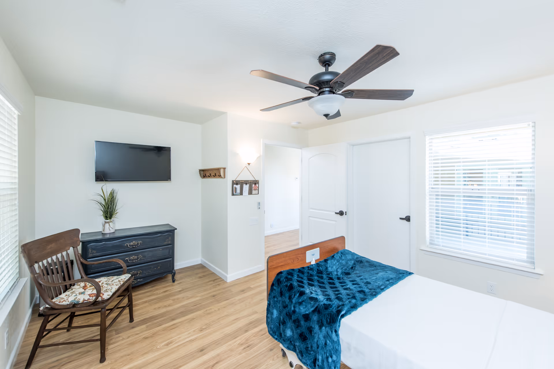 A bright bedroom with light wood flooring, a hospital-style bed with a blue blanket, a wooden chair with a floral cushion, a dark blue dresser with a plant on top, a wall-mounted flat-screen TV, a ceiling fan with dark blades, and two windows with white blinds.