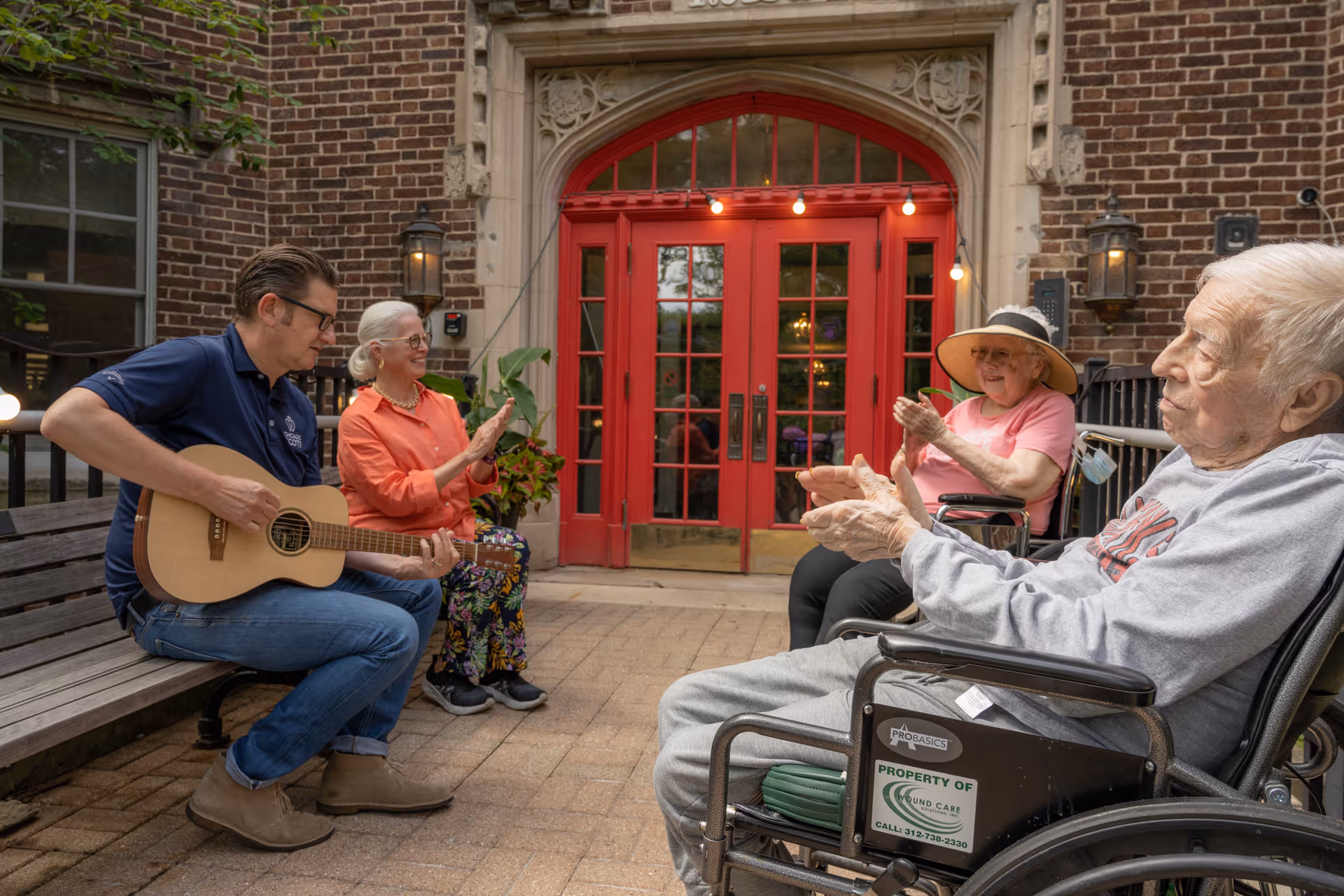 An outdoor scene at a senior living facility showing a man playing guitar while three elderly individuals, two women and one man in a wheelchair, sit nearby clapping and enjoying the music. They are seated on benches and wheelchairs in front of a building with a large red door and brick walls.