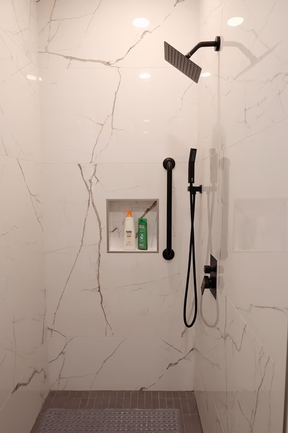 A modern shower area with white marble-patterned tiles on the walls and a gray tiled floor. The shower features a black square rain showerhead, a black handheld showerhead with hose, and a black grab bar. There is a recessed shelf in the wall holding two bottles of shower products.