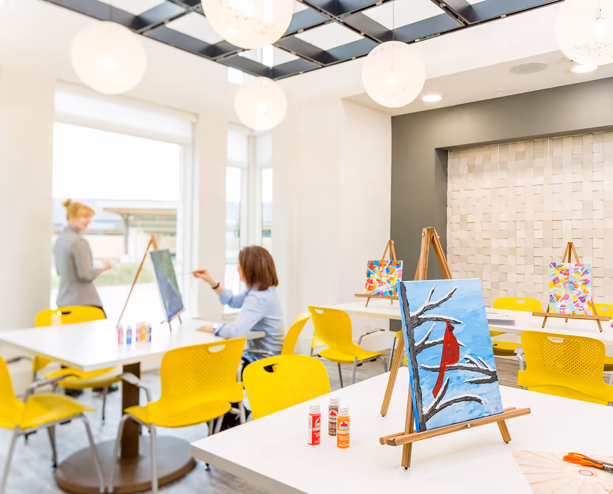 Bright activity room with easels and colorful paintings on tables, yellow chairs, and two people painting near large windows.