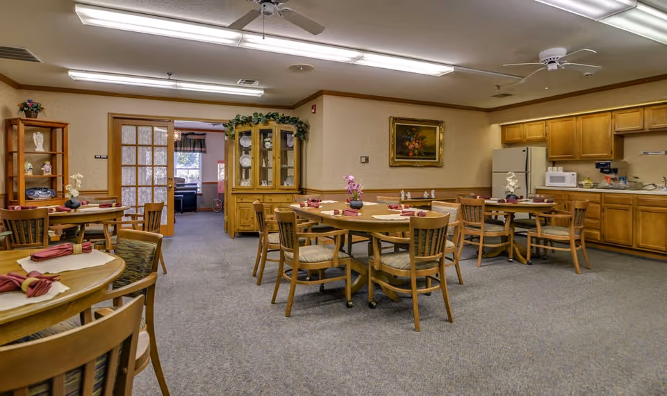 A senior living facility dining room with several round wooden tables and chairs, each table set with placemats and folded napkins. The room has carpeted floors, wooden cabinets along one wall with a refrigerator, microwave, and sink. There is a glass-front cabinet displaying dishes and a painting on the wall. Ceiling fans and fluorescent lights are mounted on the ceiling.