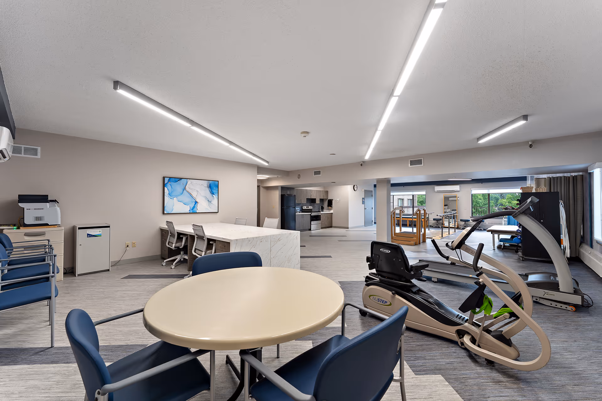 A spacious, well-lit interior room featuring a round table with four blue chairs in the foreground, exercise equipment including a recumbent bike and treadmill on the right, and a kitchen area with a refrigerator and stove in the background. The room has modern lighting, a piece of abstract blue and white artwork on the wall, and large windows letting in natural light.