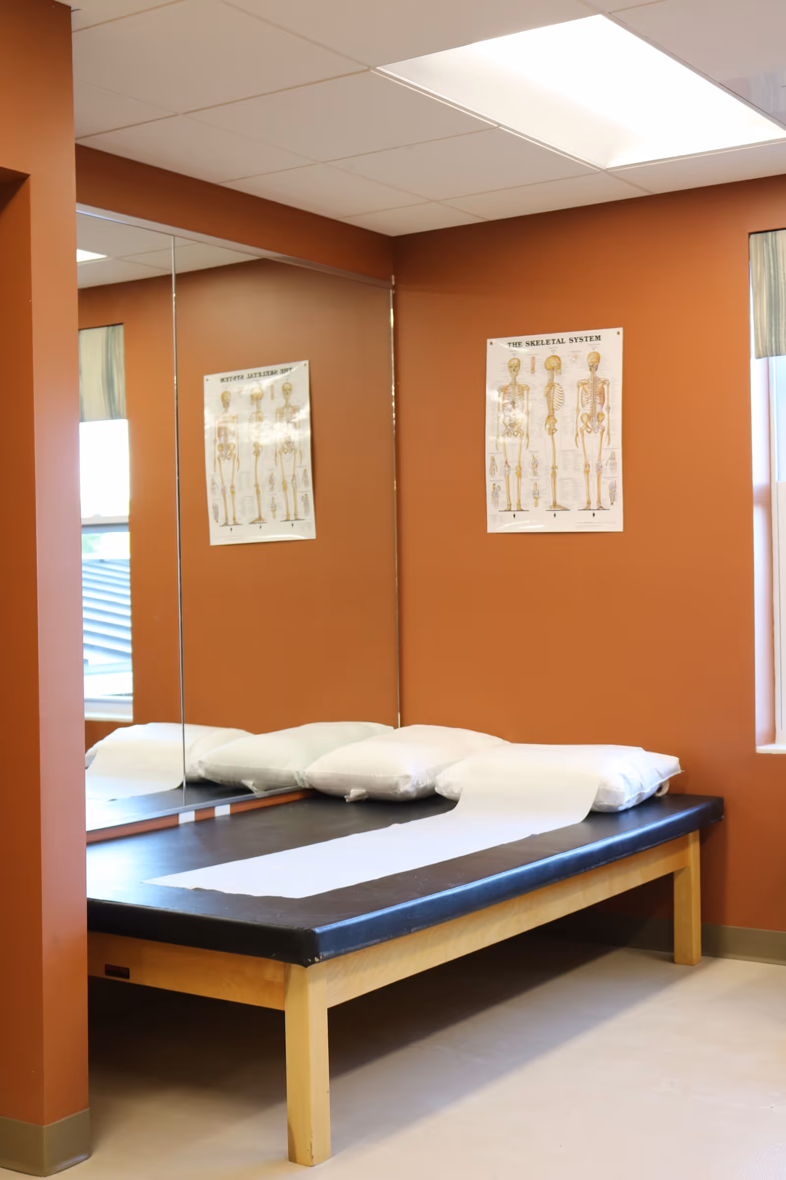 A therapy/examination room with a padded treatment table covered with paper, pillows, a wall mirror, and anatomical posters on rust-colored walls.