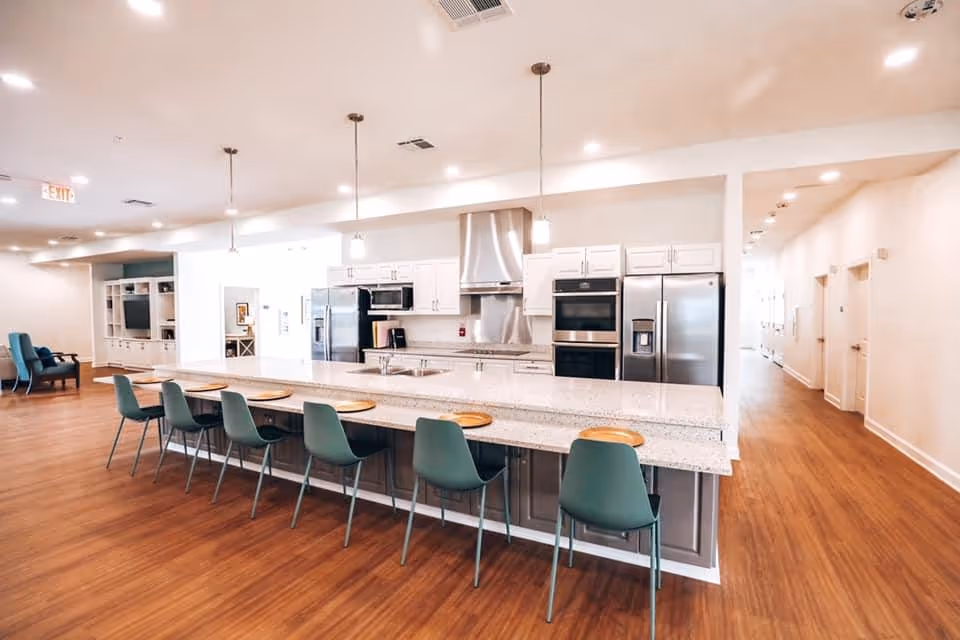 Open communal kitchen with a long island, green bar stools, and stainless-steel appliances in a bright common area.