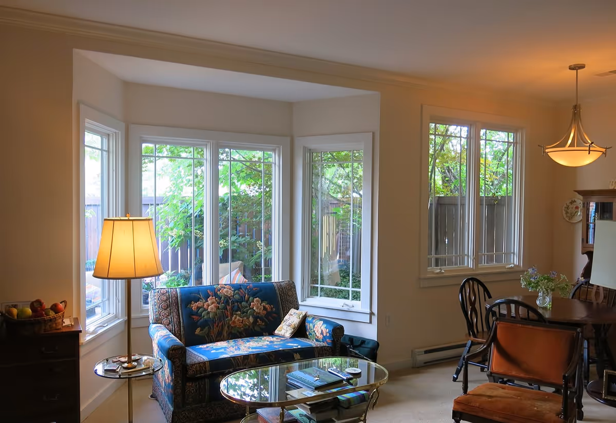 A cozy living room area with a floral patterned blue loveseat, a glass coffee table with books and remote controls, and a standing lamp beside a small side table with a basket of fruit. Large windows reveal green foliage outside. To the right, there is a dining area with a wooden table, chairs, a hanging light fixture, and a vase with flowers.