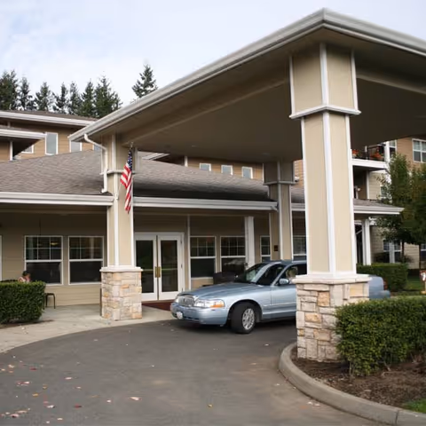 Entrance of a senior living facility with a covered driveway supported by large beige columns with stone bases. A silver car is parked under the covered area near double glass doors. An American flag is mounted on one of the columns. The building is beige with white trim and surrounded by greenery and trees in the background.